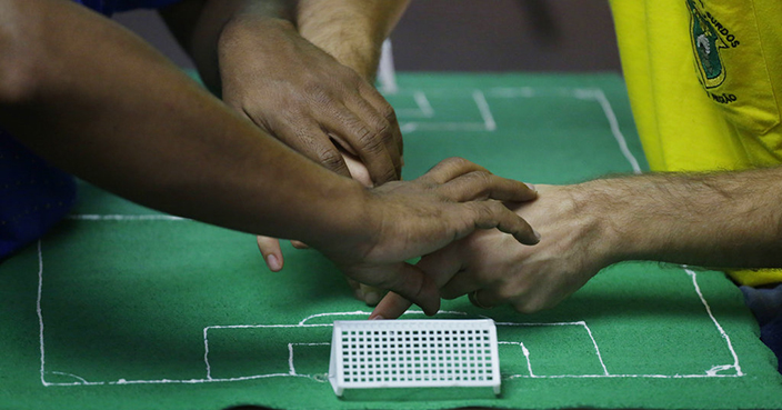 Brazil soccer fan Carlos Junior, left, who is both deaf and blind, experiences the World Cup match between Brazil and Mexico with the help of an interpreter who uses tactile signing and a model soccer field to recount the game, in Sao Paulo, Brazil, Monday, July 2, 2018. The system is this: Junior places his hands on the interpreter’s. One hand represents the ball, the other the player who has possession. The interpreter moves his hands around the model field to indicate the action. Meanwhile, another interpreter draws on Junior’s back, communicating which team and even which player (by tracing the player’s number) has the ball. The interpreter can also note fouls, yellow or red cards, blocks and saves. (AP Photo/Nelson Antoine)