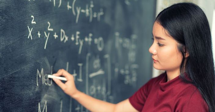 Beautiful Asian student writing on blackboard with chalk in classroom.