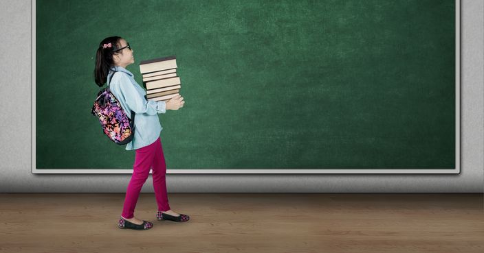 Picture of cute student carrying pile of books while walking in the classroom