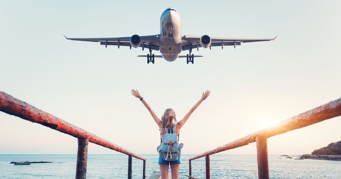 Airplane and woman at sunset. Summer landscape with girl standing on the sea pier with raised up arms and flying passenger airplane. Woman and landing commercial plane in the evening. Lifestyle