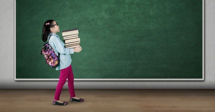 Picture of cute student carrying pile of books while walking in the classroom