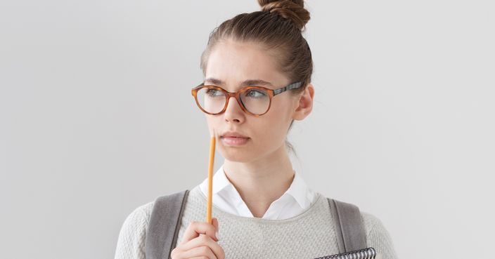Closeup portrait of good-looking young woman standing isolated on grey background, wearing glasses, holding pencil next to lips, thinking and looking leftwards, trying to find answer or solution.