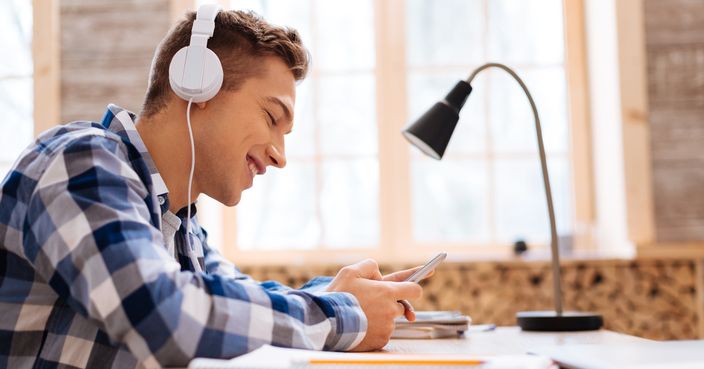 Having a rest. Good-looking content fair-haired boy smiling and wearing headphones and listening to music and holding his modern phone