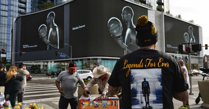 A vendor sells ice-cream as fans take photos of a giant electronic billboard featuring NBA star Kobe Bryant next to the Staples Center in downtown Los Angeles on Thursday, Jan. 30, 2020. People wearing Kobe Bryant jerseys, hats, shoes and other gear continued to arrive by the thousands at the downtown arena where Bryant had made basketball history. (AP Photo/Richard Vogel)