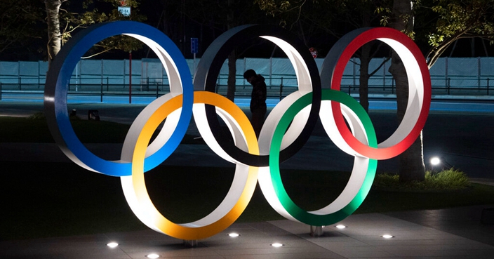 FILE - In this Tuesday, March 24, 2020 file photo, a man is seen through the Olympic rings in front of the New National Stadium in Tokyo. The Tokyo Olympics were postponed a month ago. But there are still more questions than answers about the new opening on July 23, 2021 and what form those games will take.(AP Photo/Jae C. Hong, File)